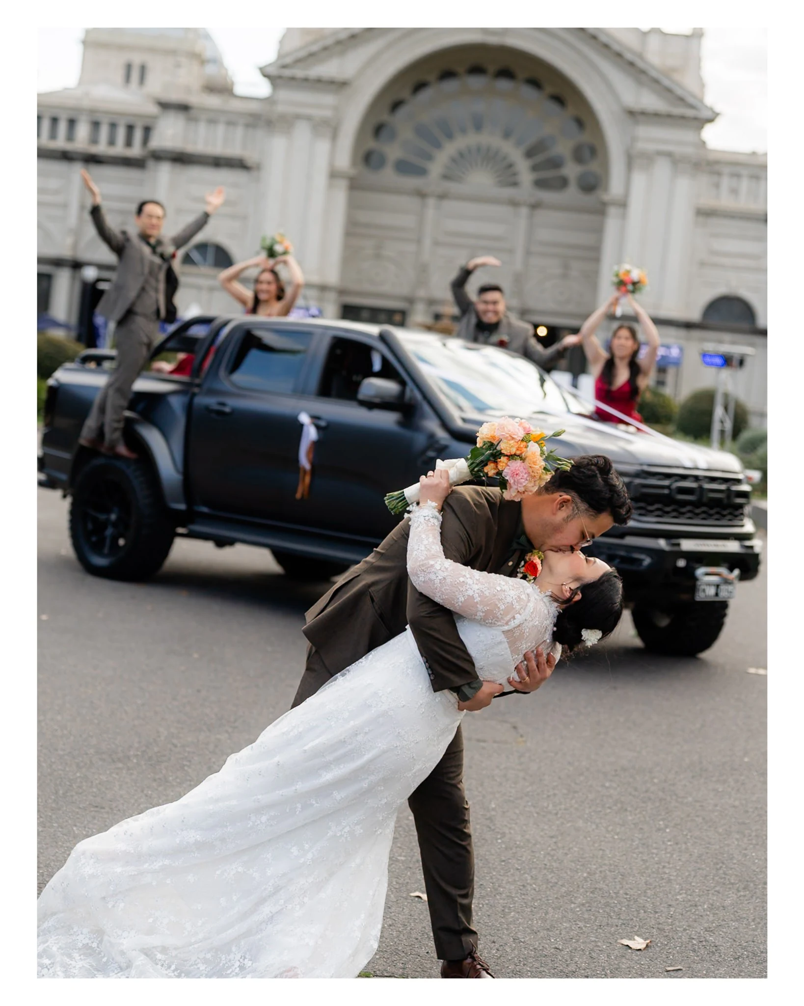 Newlyweds sharing a dip kiss beside their wedding car and cheering wedding party.