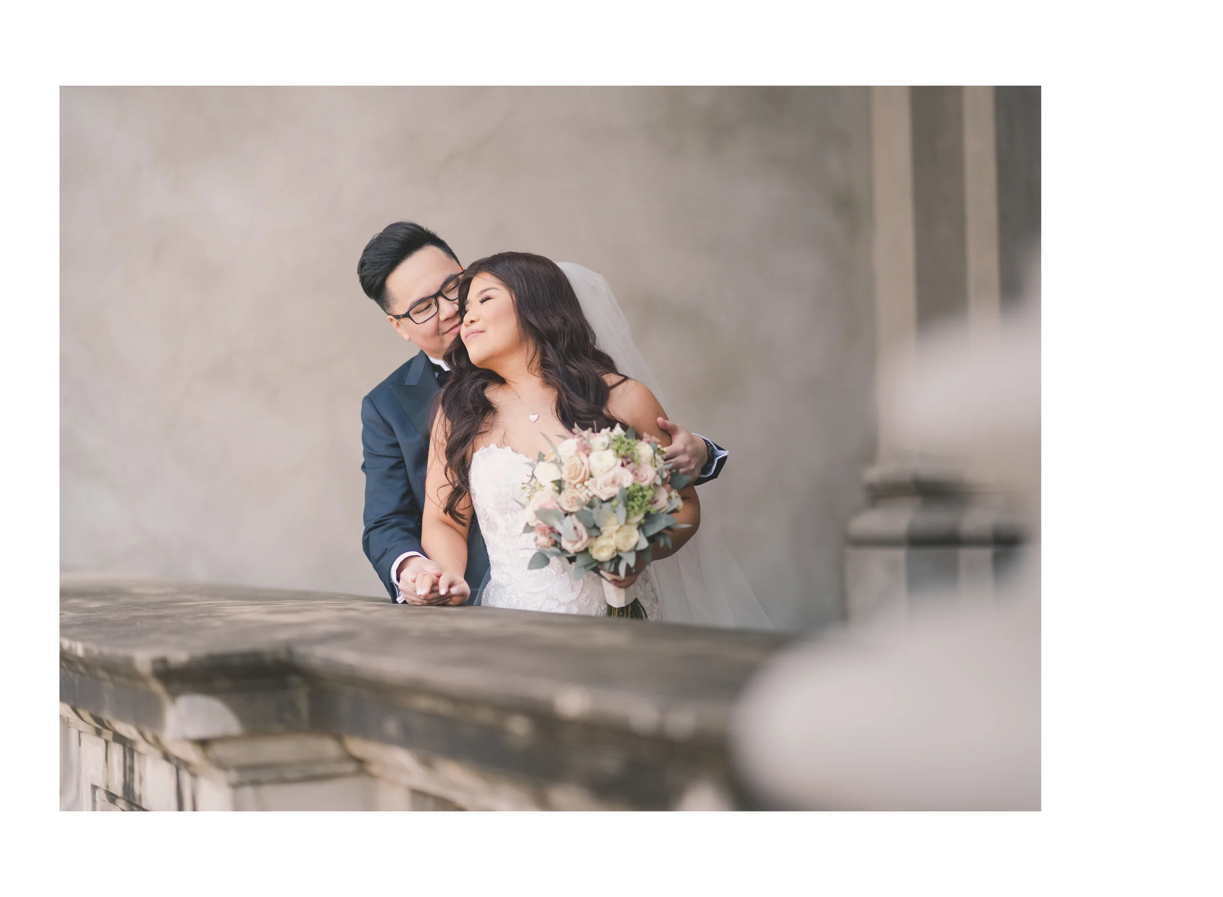 Newlyweds sharing a quiet embrace behind a stone balustrade, the bride holding a blush bouquet