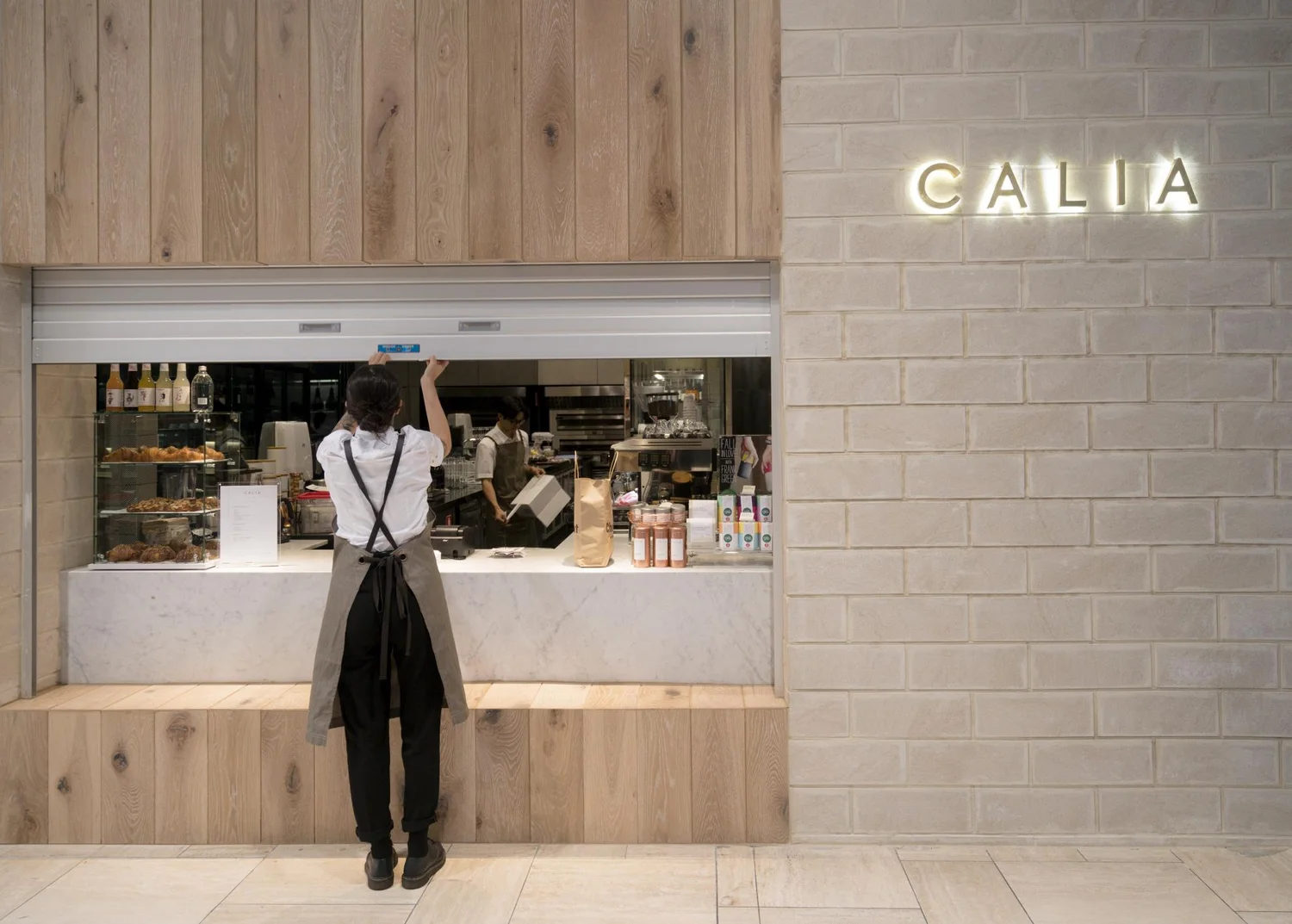 Person standing at the pale timber Calia counter beneath its illuminated sign